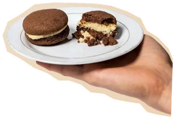 Tangan memegang piring putih berisi dua sandwich cookie KriWul, salah satunya terbelah memperlihatkan isian.