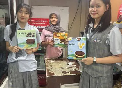 Dua siswi tim KriWul berfoto bersama pemilik toko oleh-oleh sambil memegang kotak KriWul.