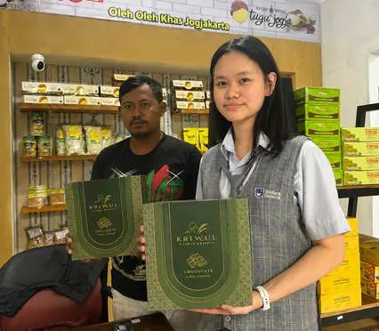 Siswi tim KriWul memegang kotak KriWul di depan rak toko bersama penjaga toko.