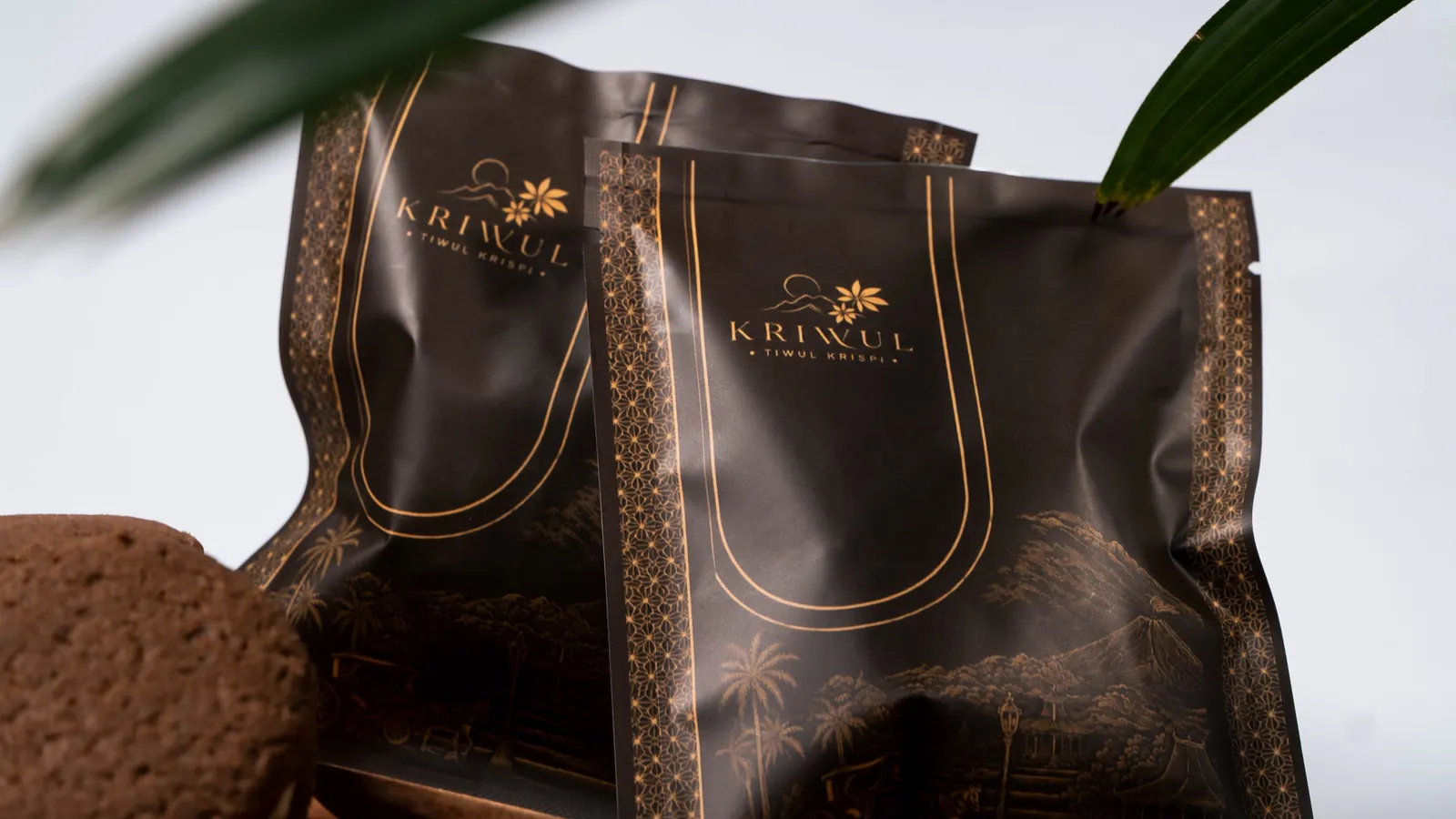 Dua pouch KriWul cokelat tua berilustrasi emas di samping daun palem, dengan sekeping kukis di depannya.