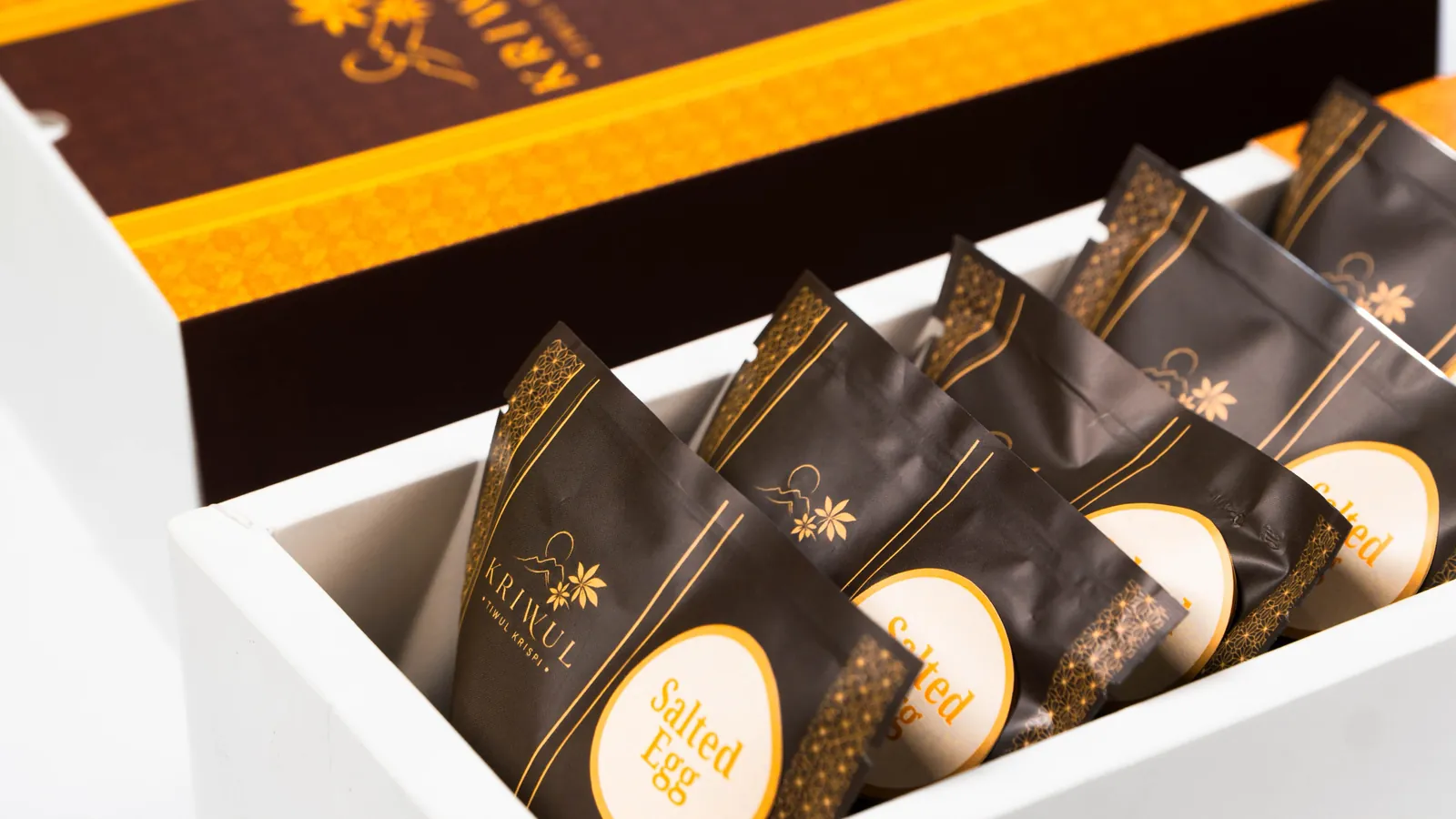 Deretan pouch KriWul cokelat tua berlabel bundar krem bertuliskan Salted Egg di dalam baki putih, dengan kotak oranye di belakangnya.